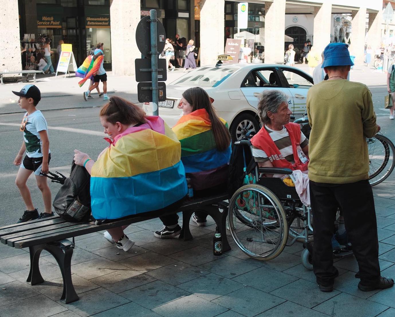 People wearing rainbow flags as capes sitting on bench